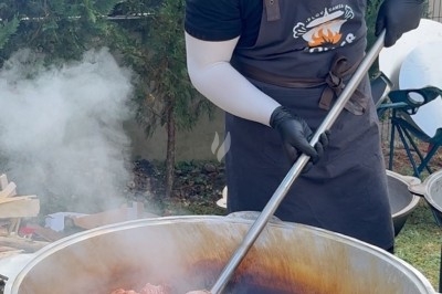 Homme en tablier cuisant de gros morceaux de viande dans une grande marmite fumante en extérieur.