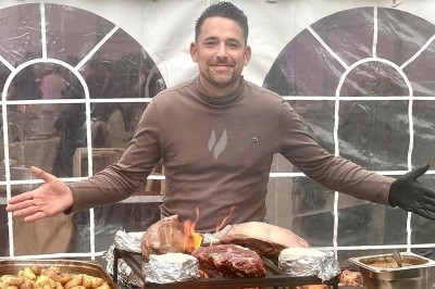 Homme souriant devant un barbecue chargé de viandes et pommes de terre, sous une tente blanche.