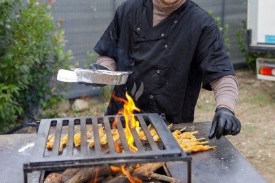 Un cuisinier prépare des brochettes de viande sur un barbecue en plein air avec des flammes vives.
