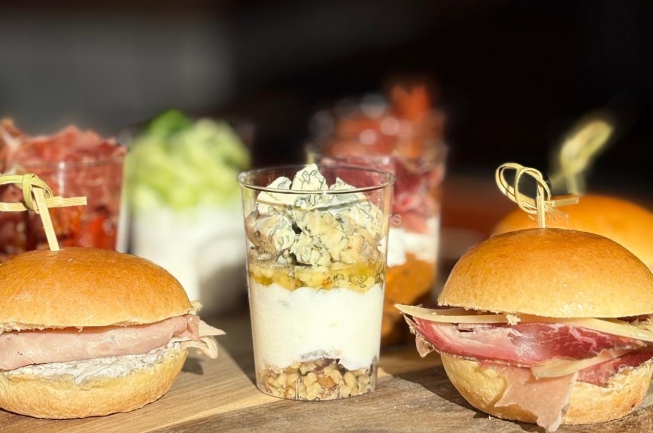 Assortiment de mini-burgers au jambon et verrines garnies de fromage, noix et légumes sur une planche en bois.