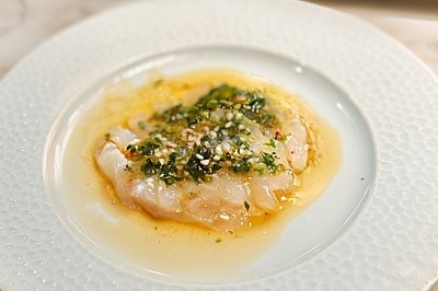 Carpaccio de poisson blanc finement tranché, assaisonné d'herbes, d'épices et d'huile, servi sur une assiette blanche.