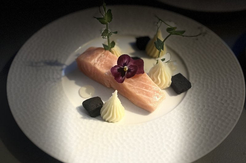 Traiteur La Petite Cuillère Traiteur Theix #2 Pavé de saumon rosé dressé sur une assiette blanche texturée, accompagné de quenelles crémeuses, de petits cubes noirs et d'une fleur violette.