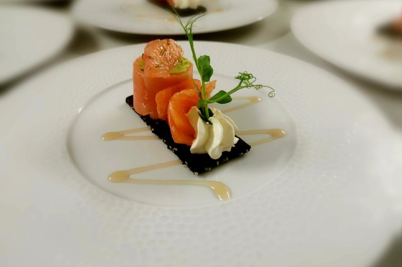 Traiteur La Petite Cuillère Traiteur Theix #3 Bouchée de saumon roulé sur un fond noir sésame, accompagnée d'une crème blanche, d'une pousse verte et d'un filet de sauce beige sur assiette blanche.