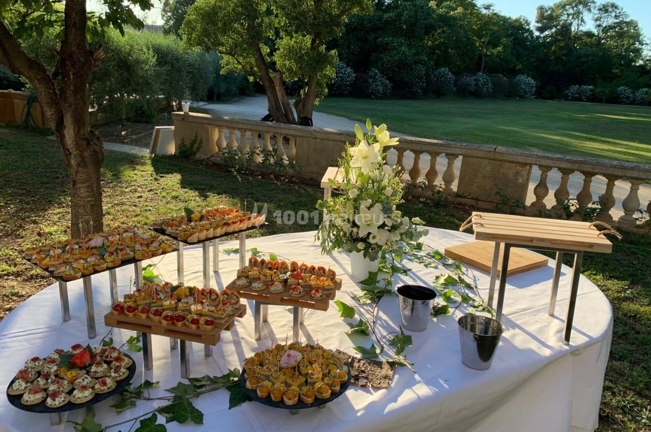 Traiteur Les Sens du Lez Saint-Jean-de-Védas #1 Buffet en extérieur avec des amuse-bouches variés disposés sur des présentoirs, entouré de verdure et de fleurs.